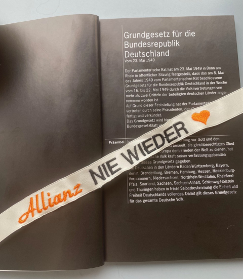 Auf dem Grundgesetz der Bundesrepublik Deutschland liegt ein Band auf dem "Allianz NIE WIEDER ♥" geschrieben steht.